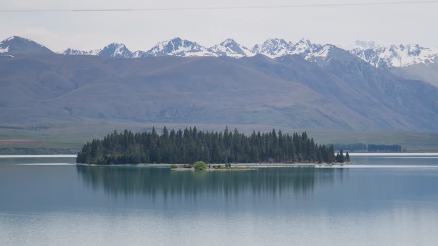 A static, zoomed in shot of Matuariki island in the middle of Lake Tekapo, New Zealand with snowy mountains in the background.