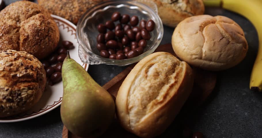 A warm tabletop slider shows crusty buns on a wooden plate beside pears, bananas, and a small bowl of nuts in chocolate. 