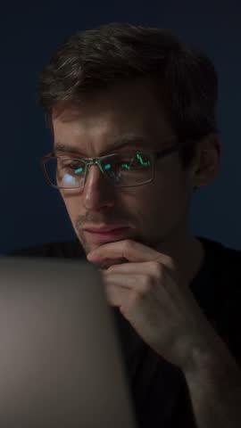 A smiling crypto trader wearing glasses clenches fists in excitement while looking at stock market charts reflected in his lenses on a laptop screen. Happy trader celebrating stock market success. 