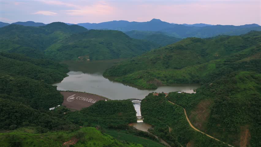 Aerial dolly in over mountain valley approaching El Carrizo Dam and reservoir, with CONAGUA hillside lettering visible in Jalisco, Mexico