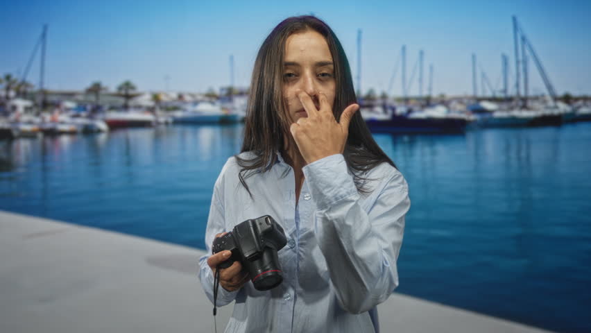 Woman holding camera and showing v sign with fingers on street by marina boats and pier; confidence focus.