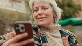 Smiling senior woman using a smartphone outdoors. She's wearing a beige turtleneck and a plaid jacket. Slow-motion. - Powered by Shutterstock - Get 15% off with code: PIKWIZARD15