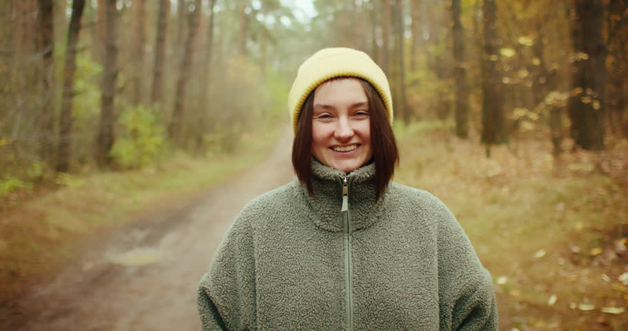 Close-up portrait of a happy young woman in a yellow beanie smiling at the camera in an autumn forest