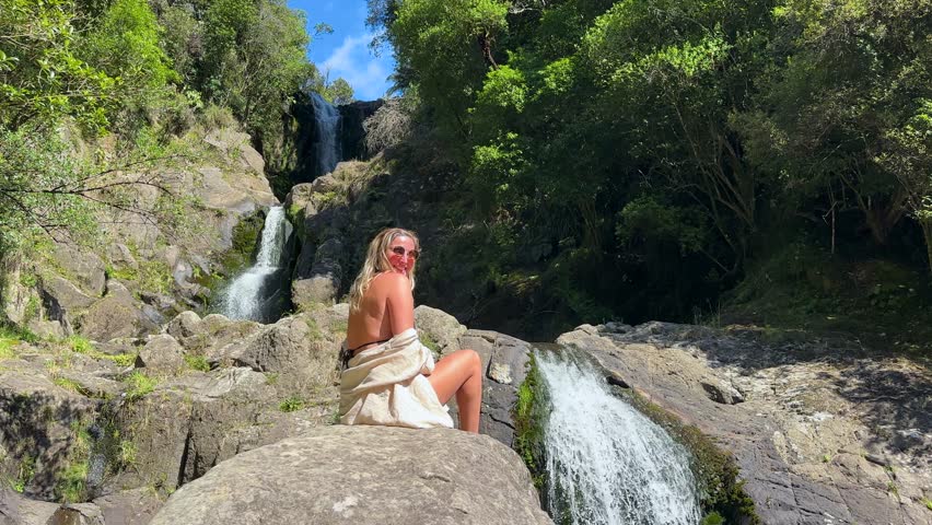 A woman sits on a rock admiring the stunning tiered Kaiate Falls in New Zealand. Surrounded by lush forest, the cascading water creates a peaceful and natural scene in the heart of nature.