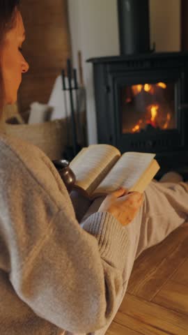 Woman sitting with hot drink by the burning fireplace and reading a book