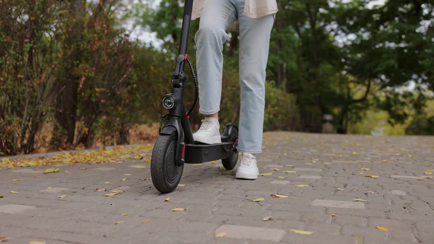 Close up cropped photo shot of young woman she wearing blue jeans denim pants riding modern electric scooter resting relaxing in fall city park go down alley outdoors on nature. Urban leisure concept