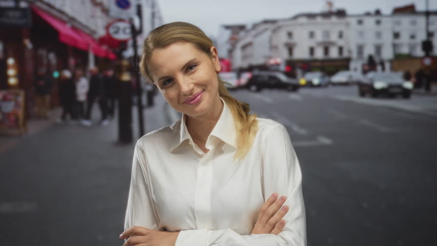 Woman smiling confidently in a city street with arms crossed wearing a white shirt outdoors in an urban european setting with traffic and pedestrians in the background.