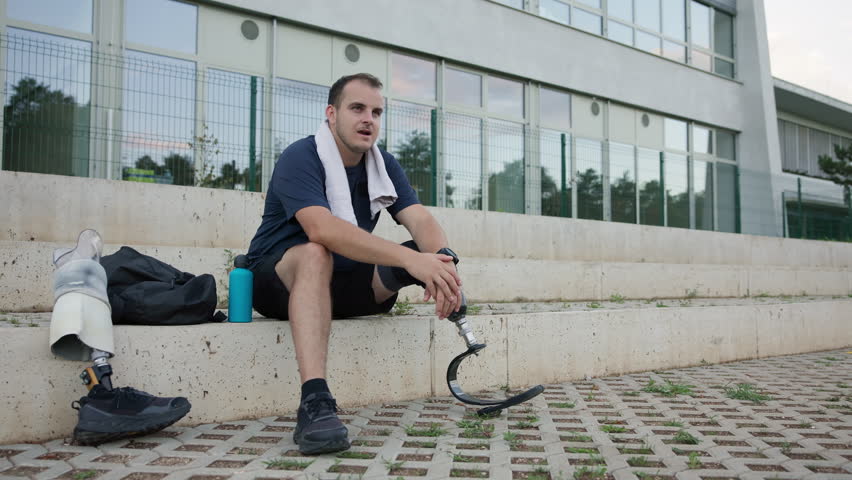 Male athlete with a disability wearing a running blade, sitting by a prosthetic leg with a sneaker, and hydrating after training, static shot. Active lifestyle concept.