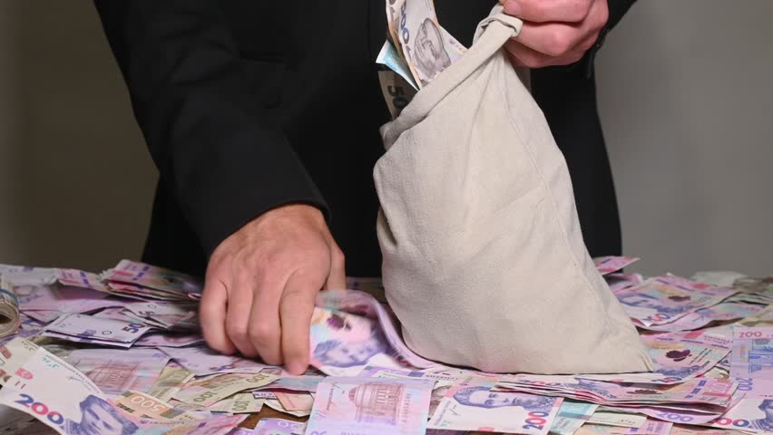 man hurriedly fills bag with stacks of Ukrainian hryvnia, sack full of money representing economic crime and illicit profit. concepts of bribery, theft and hiding illegal cash in Ukraine.