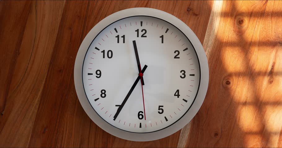 Time lapse of rotating hour, minute and second hands of a white face analog wall clock at high noon. Close up shot, sunlight and window shade passing through the frame, no people