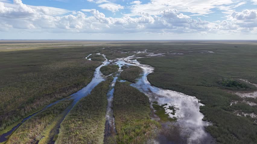 Aerial view of winding water channels in the Everglades National Park, Florida, reflecting the sun and clouds on the calm surface. The mirror-like water creates a surreal and peaceful landscape surrou