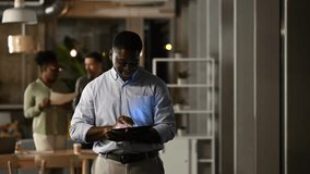 Focused african american businessman standing and using a digital tablet in a contemporary office, working late with colleagues in the background - Powered by Shutterstock - Get 15% off with code: PIKWIZARD15