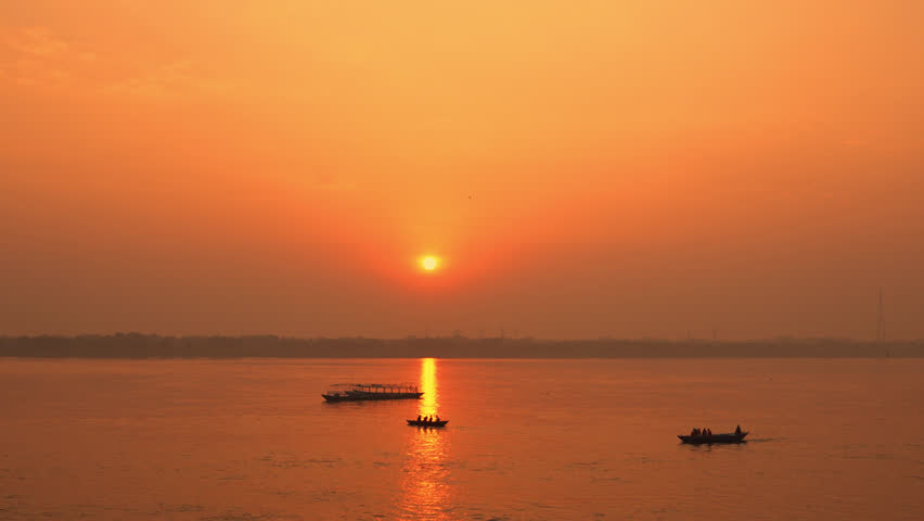 Beautiful time-lapse of the sunrise over the sacred Ganga River in Varanasi. The city slowly awakens under a golden-orange sky as boats drift across the calm waters.