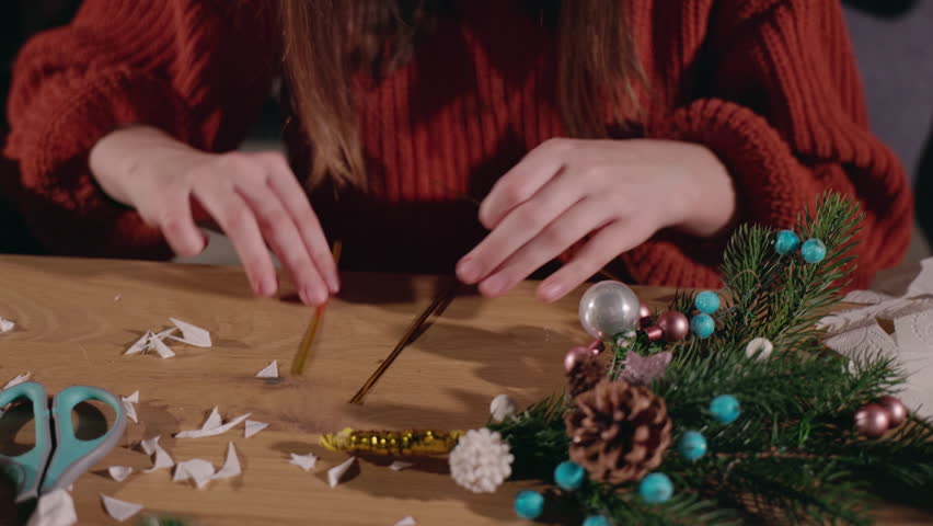 Close-up of a person folding paper for handmade Christmas decorations at a wooden table with festive branches, pinecones, and colorful ornaments. High quality 4k footage