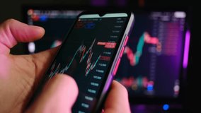 Close up of a trader hand holding a smartphone, analyzing the bitcoin price chart for investment purposes with a financial graph on a computer monitor visible in the background - Powered by Shutterstock - Get 15% off with code: PIKWIZARD15