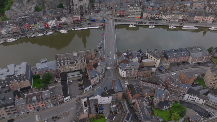 Cars drive on the Charles De Gaulle bridge in Dinant, Belgium. Boats dock along the Meuse riverbanks. Tourists visit this popular spot
