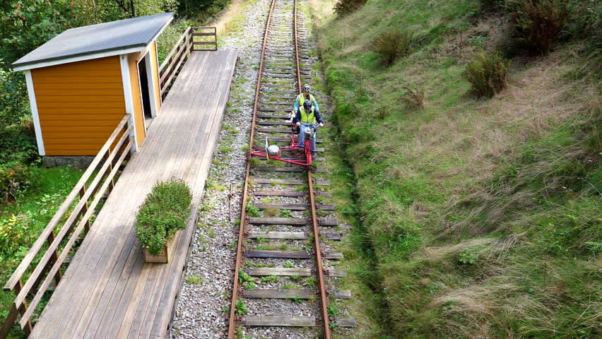 Young couple wearing high visibility safety vests and cycling helmets riding a dresine, dresin, draisine, drezine or rail bike past wooden train stop on railway in Norway, high angle view.