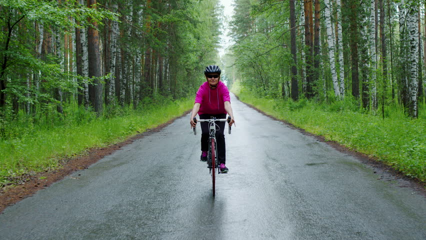 Active mature woman enjoying a bike ride on a wet asphalt road through a lush green forest, wearing a protective helmet and pink sportswear for her outdoor cardiovascular exercise