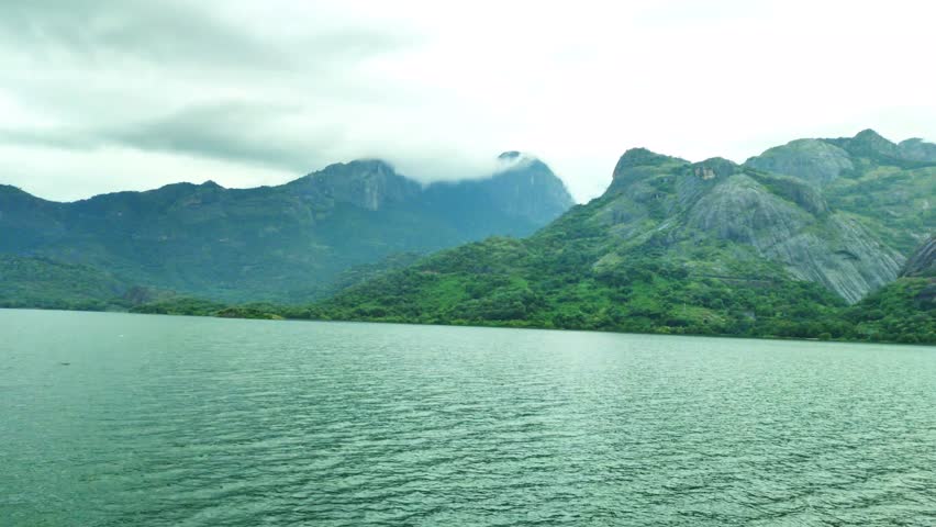 A lake at Aliyar Dam Reservoir in Tamil Nadu, India, framed by green hills and rocky peaks under a cloudy sky. Ideal for nature, travel, and outdoor landscape themes, conveying calm open wilderness.