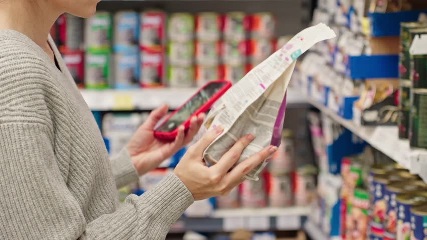 Woman scanning dry pet food using smartphone in pet store. Modern customer checking product information and ingredients using mobile app, AI assistant. Smart shopping Conscious choice Digital retail
