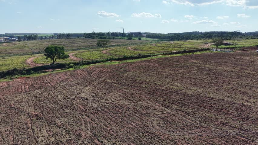 A scenic aerial perspective reveals a vast agricultural landscape, featuring a tilled field, a distant factory, and scattered trees beneath a partly cloudy sky.