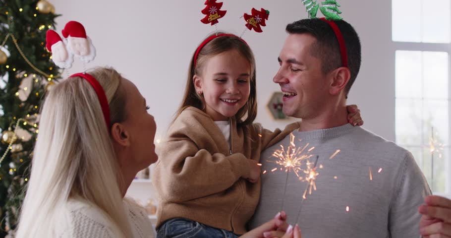 Christmas family with daughter holding sparklers indoors. At home by festive tree, smiling parents hug child girl, enjoying warm lights and cozy sparkle. Celebration, love and winter holiday joy.