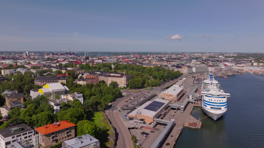 Aerial perspective of a cruise ship docking at a bustling port in Helsinki, Finland. The cityscape stretches out under a clear summer sky