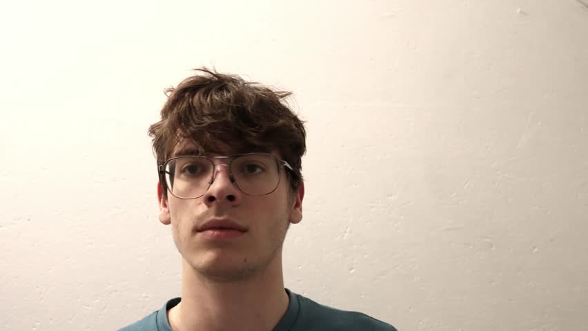 A friendly, smiling young man with glasses waves hello or goodbye at the camera. Welcoming greeting gesture.