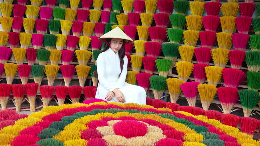 4K slow motion video of Asian woman wearing white Vietnamese cultural dress with conical hat sitting among colorful incense sticks.