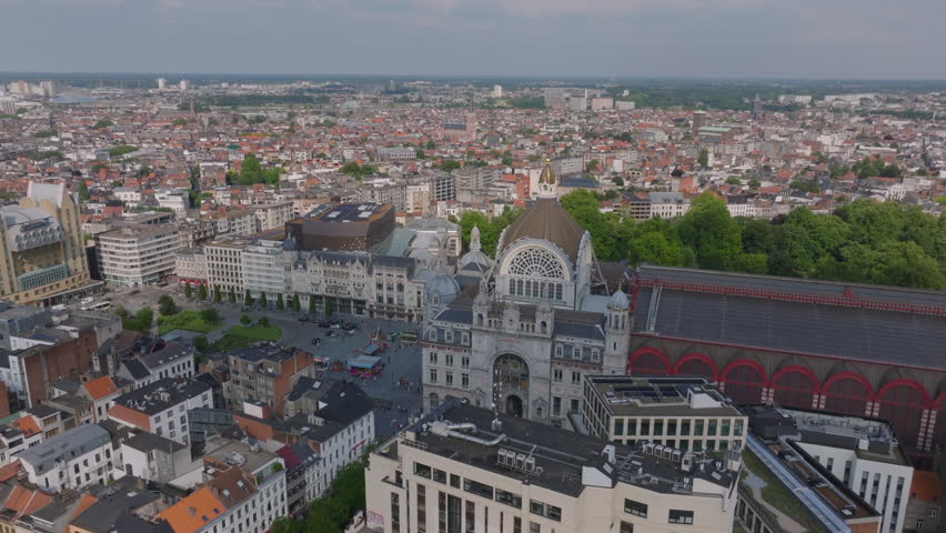 Antwerp Central Station with a dome and spire is surrounded by a busy city center with cars driving on roads and pedestrians walking on sidewalks