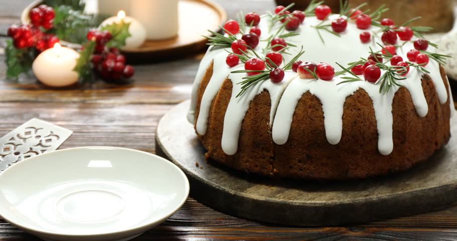 Woman taking and putting piece of delicious Christmas cake with icing, cranberries and rosemary onto plate at wooden table, closeup