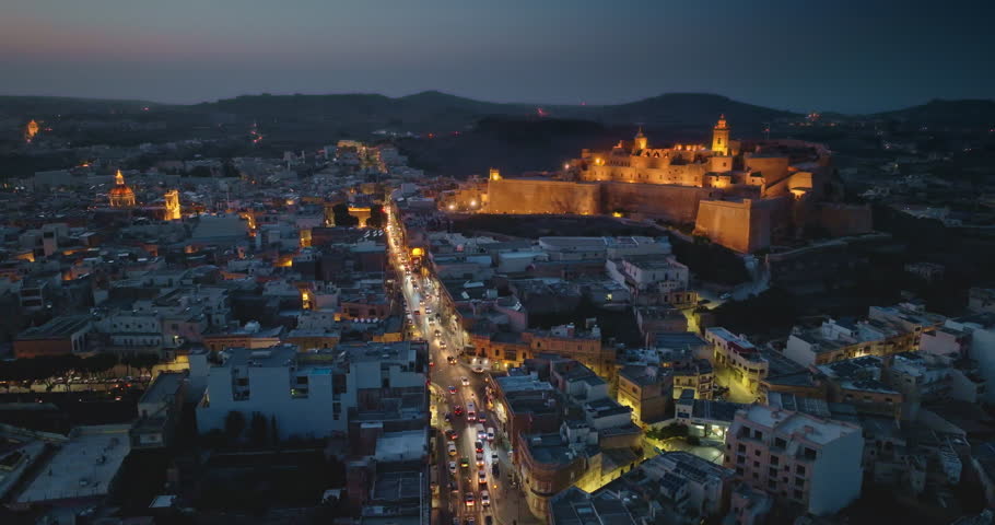 Malta, Comino island: Aerial view illuminated historic fortified Cittadella. Modern city buildings and streets glowing over Victoria on Gozo during enchanting twilight hours. Drone flight footage