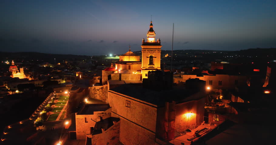 Malta, Comino island: Aerial view illuminated Citadel of Victoria glowing at dusk, highlighting its historic architecture and the surrounding Gozo cityscape at night. Drone flight footage panorama