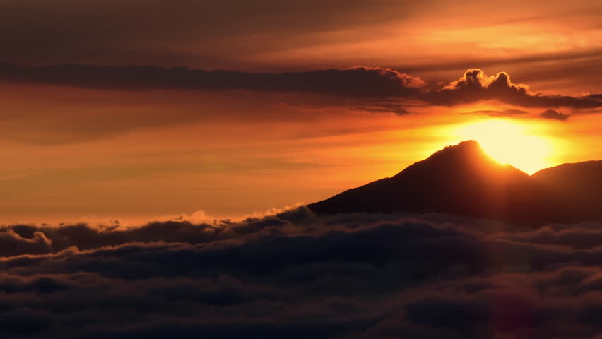 Bali, Mount Batur volcano silhouette rising above soft clouds layer during vibrant golden and orange sunset, creating serene and breathtaking natural landscape. Nature background. Aerial view panorama