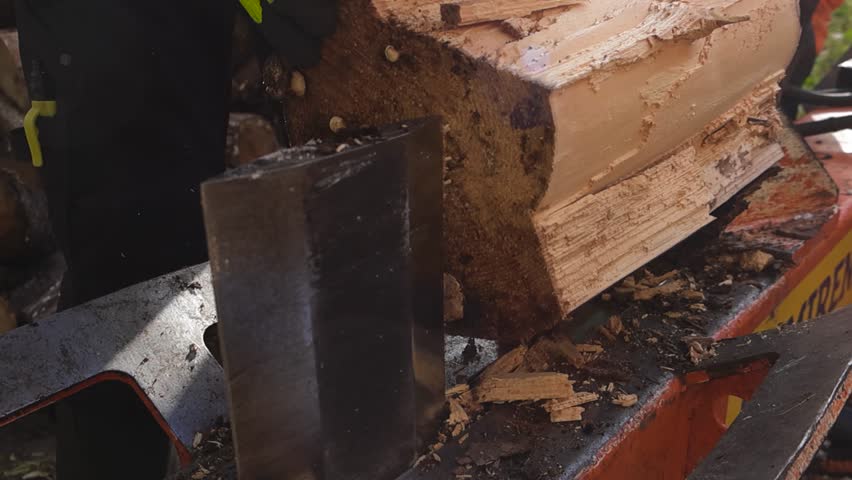 Large wooden log being split by a wood splitter machine in slow motion during a sunny autumn day with worker with gloves helping it to stand still. Sawdust and woodchips flying around in the sunshine.