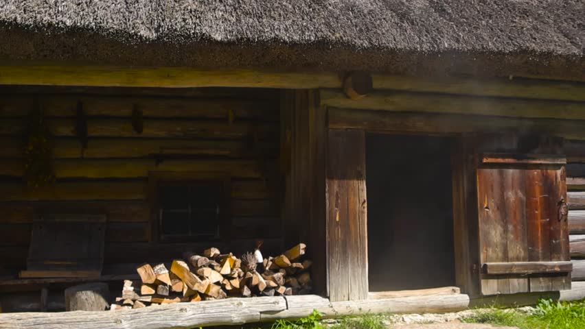 Low angle slow pan of traditional smoke sauna, ancient log house. Barn dwelling facade with thatched roof and stacked firewood. Open chamber wooden door releasing stream of smoke into the summer air