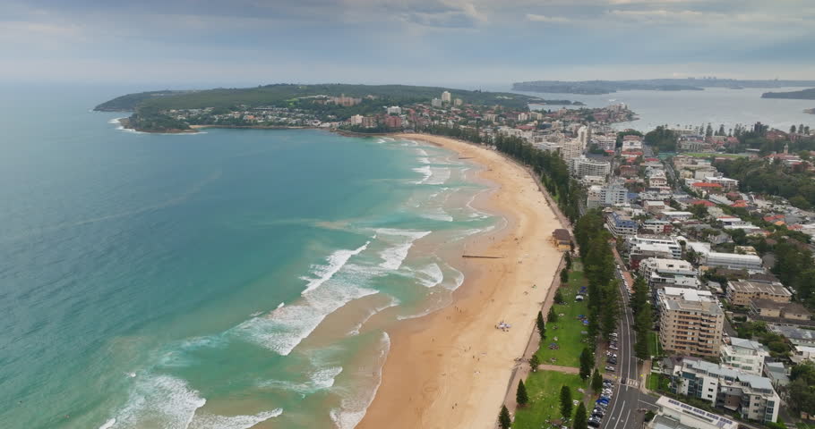 Manly Beach in Sydney, Australia, coastline with golden sand and turquoise ocean waves breaking the shore, city urban development and residential buildings under bright sky. Aerial view drone footage