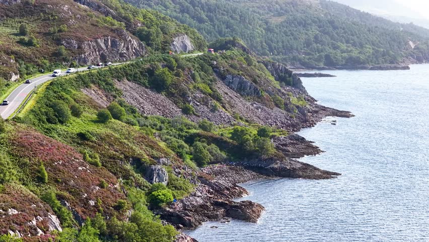 Vehicles travel on winding coastal road beside cliffs and ocean, bright daylight, smooth aerial movement