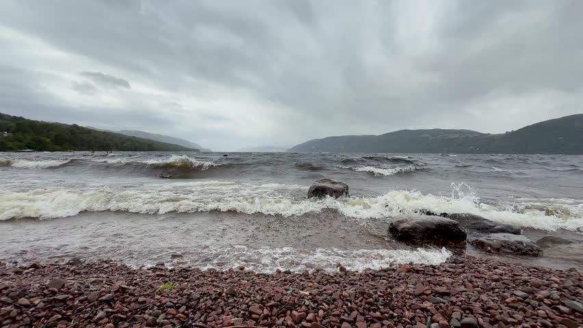 Strong waves hit rocky Loch Ness beach under overcast skies, wide shot, static camera, daytime