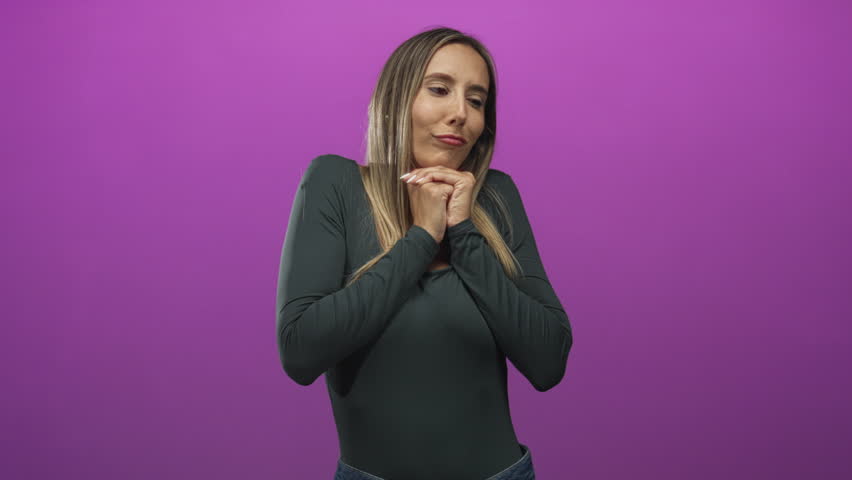 Woman clasping hands and leaning slightly forward in studio with magenta backdrop wearing dark green top and jeans; hopeful longing.