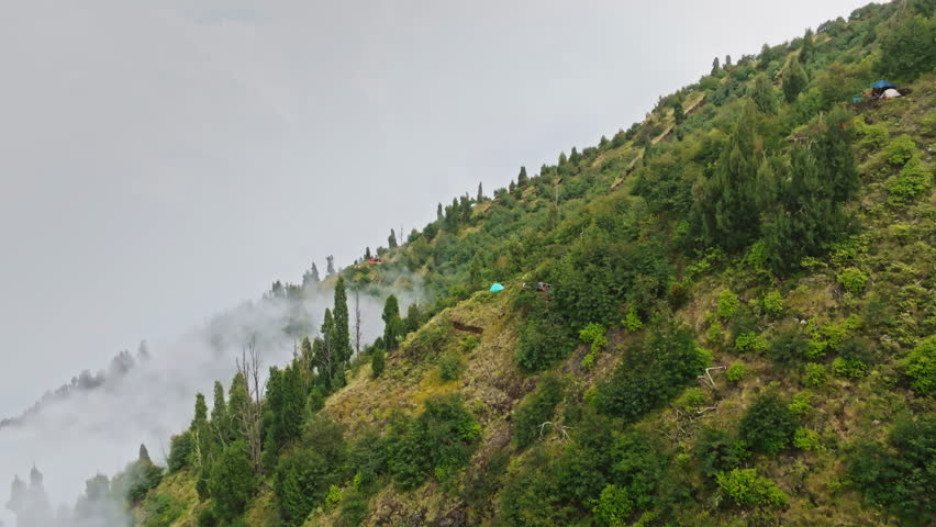 Mount Agung volcano slope in Bali featuring hikers on a trekking path and small camping tents nestled among green trees, with atmospheric fog moving through the lush landscape