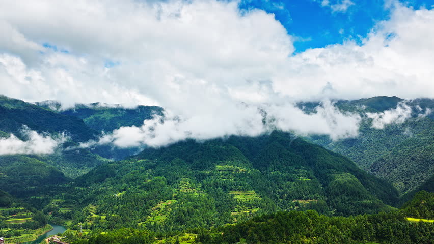 Aerial shot of the beautiful green mountain range with lush forest landscape covered by clouds and mist under a blue sky