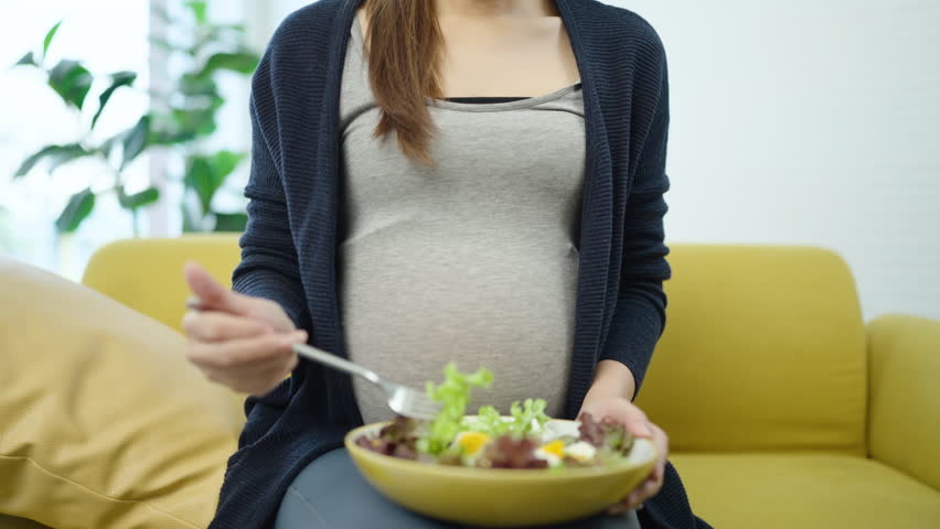 Expectant mother enjoying fresh vegetables for baby nourishment, symbol of healthy pregnancy and balanced diet. Pregnant woman eating healthy green salad on sofa.
