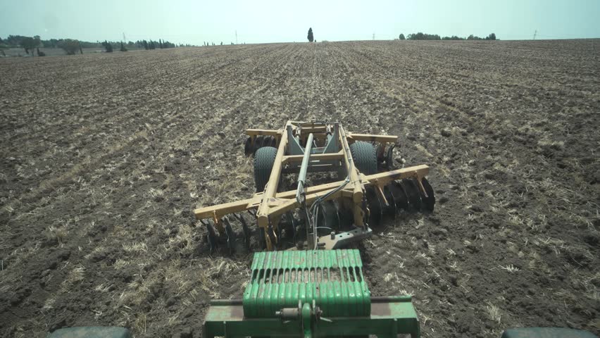 Time Lapse of a disc harrow pulled behind a tractor, shot from the rear window. Preparing the field for sowing, dust rising from the discs under bright summer skies. Driver