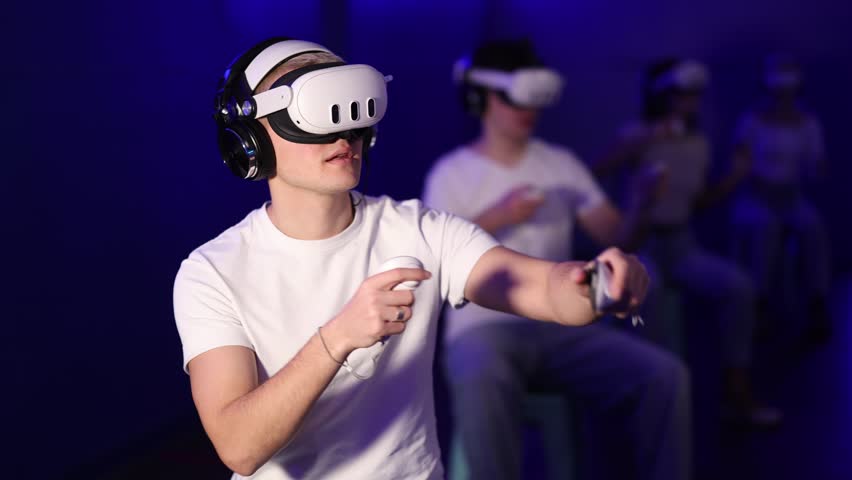 Young guy in a VR helmet holds two joysticks in his hands and plays video games in virtual reality with friends. Gamer guy in headphones and a virtual reality helmet immersed in a video game - Powered by Shutterstock - Get 15% off with code: PIKWIZARD15