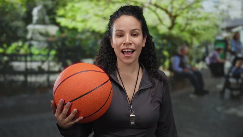 Woman holding basketball in park setting with bright greenery, expressing enthusiasm and joy, suggesting outdoor sports coaching or recreation activities.