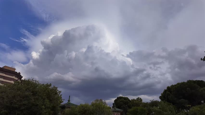 Timelapse of a massive storm cloud evolving in a blue sky over some buildings and trees. Low-angle shot.
