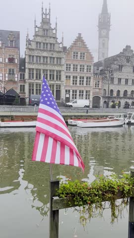 A foggy morning in a quaint town Ghent with historic architecture. An American flag stands proudly in front of the serene river, surrounded by beautiful buildings.