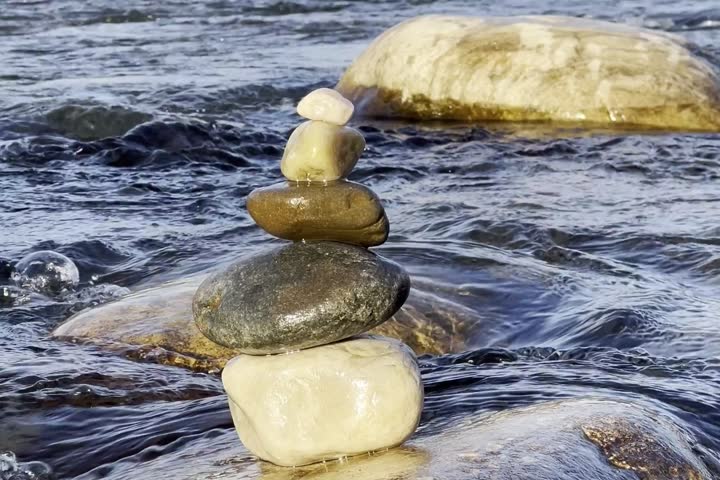 A close-up shot of a stack of smooth river rocks balanced in the middle of a flowing river, symbolizing balance, mindfulness, and tranquility. Water rushes around the stones under the sunlight.