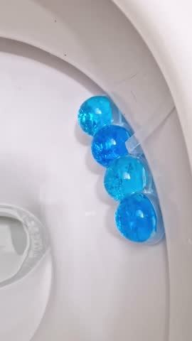 Vertical, close-up shot of a white ceramic toilet bowl flushing. Vibrant blue gel cleaner releases into the clear water, creating suds for hygiene and freshness in a modern bathroom environment.